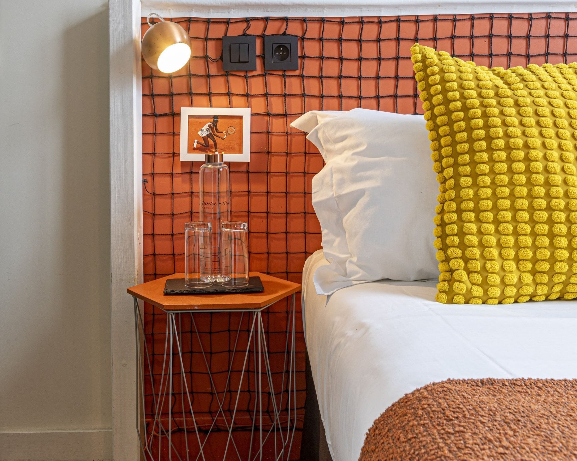 Détail de la chambre de l’Hôtel Olympic à Boulogne-Billancourt avec tête de lit inspirée d’un filet de tennis, lampe de lecture et bouteille d’accueil.