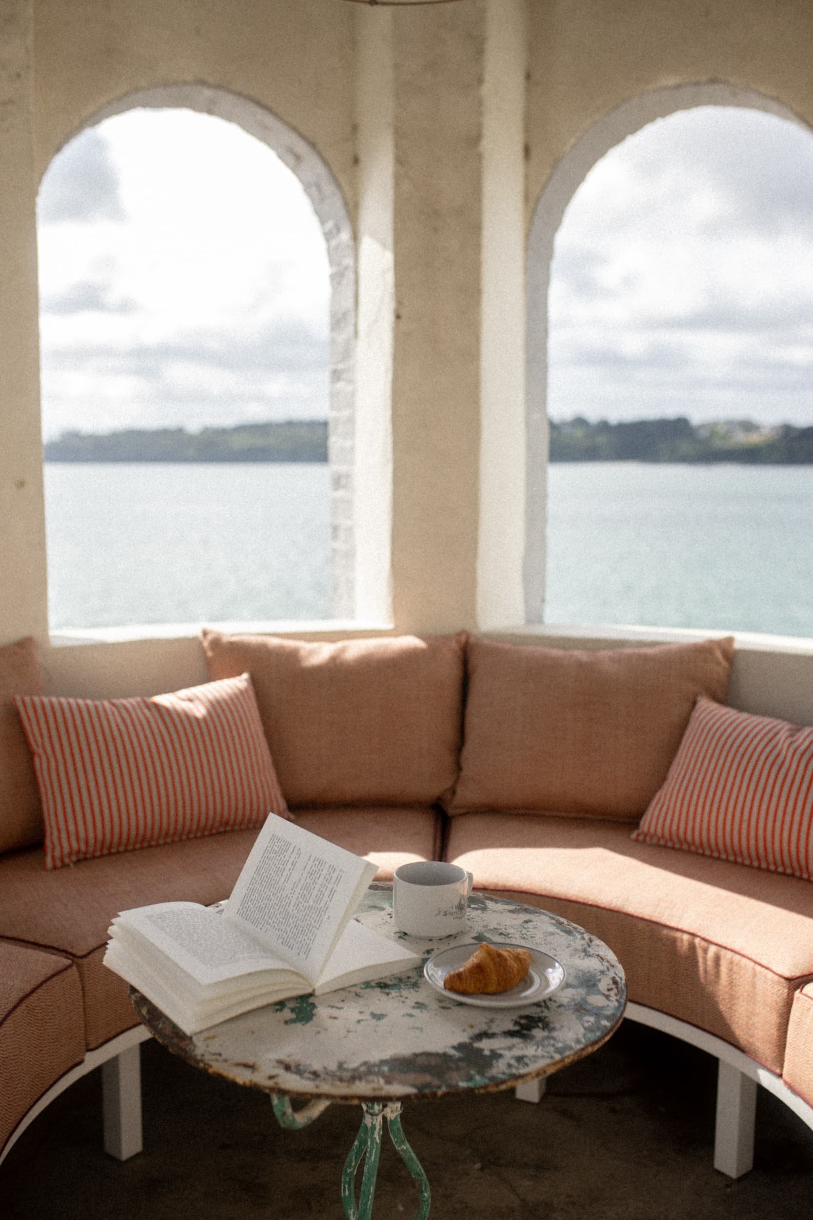 Coin salon avec vue sur la mer, un livre ouvert, une tasse de café et un croissant sur une table.