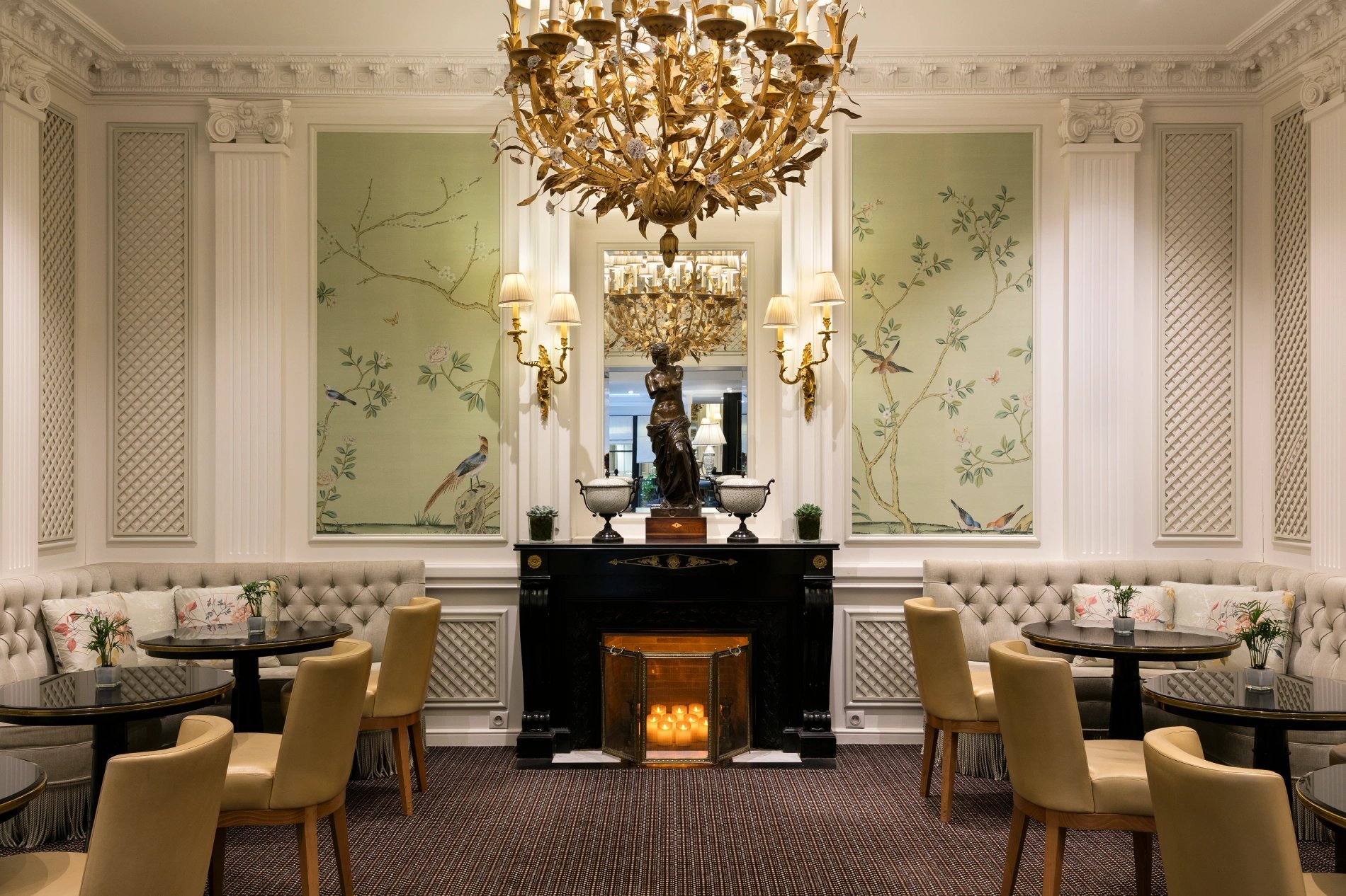 Salon élégant d'un hôtel avec un lustre doré, des tables et des chaises autour d'une cheminée.