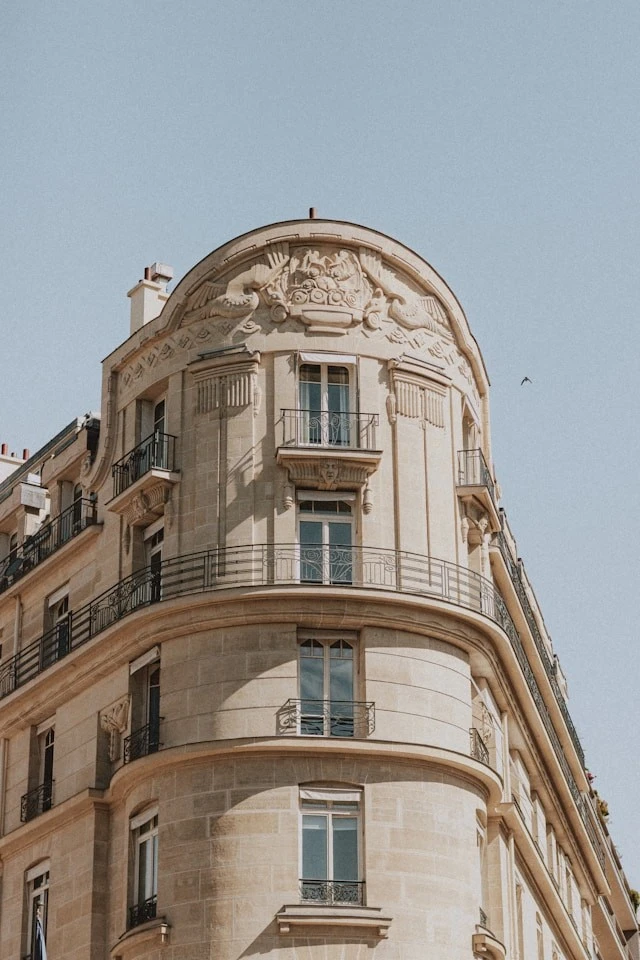 Makom Ferdinand Duval, Façade haussmannienne avec des balcons en fer forgé et des sculptures ornementales, sous un ciel bleu clair.