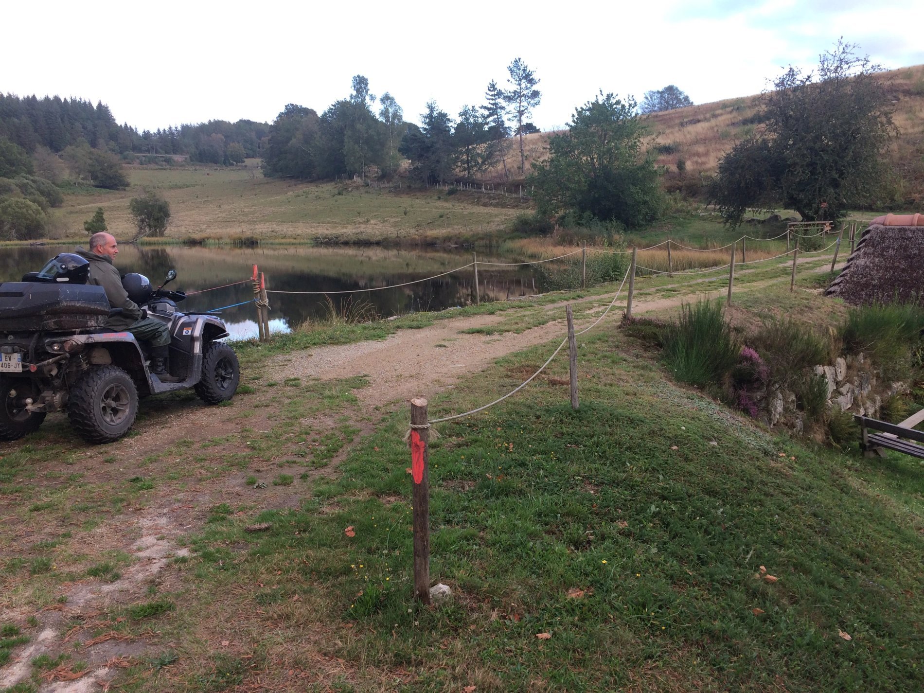 Homme en quad roulant sur un chemin de terre, longeant un petit étang clôturé.