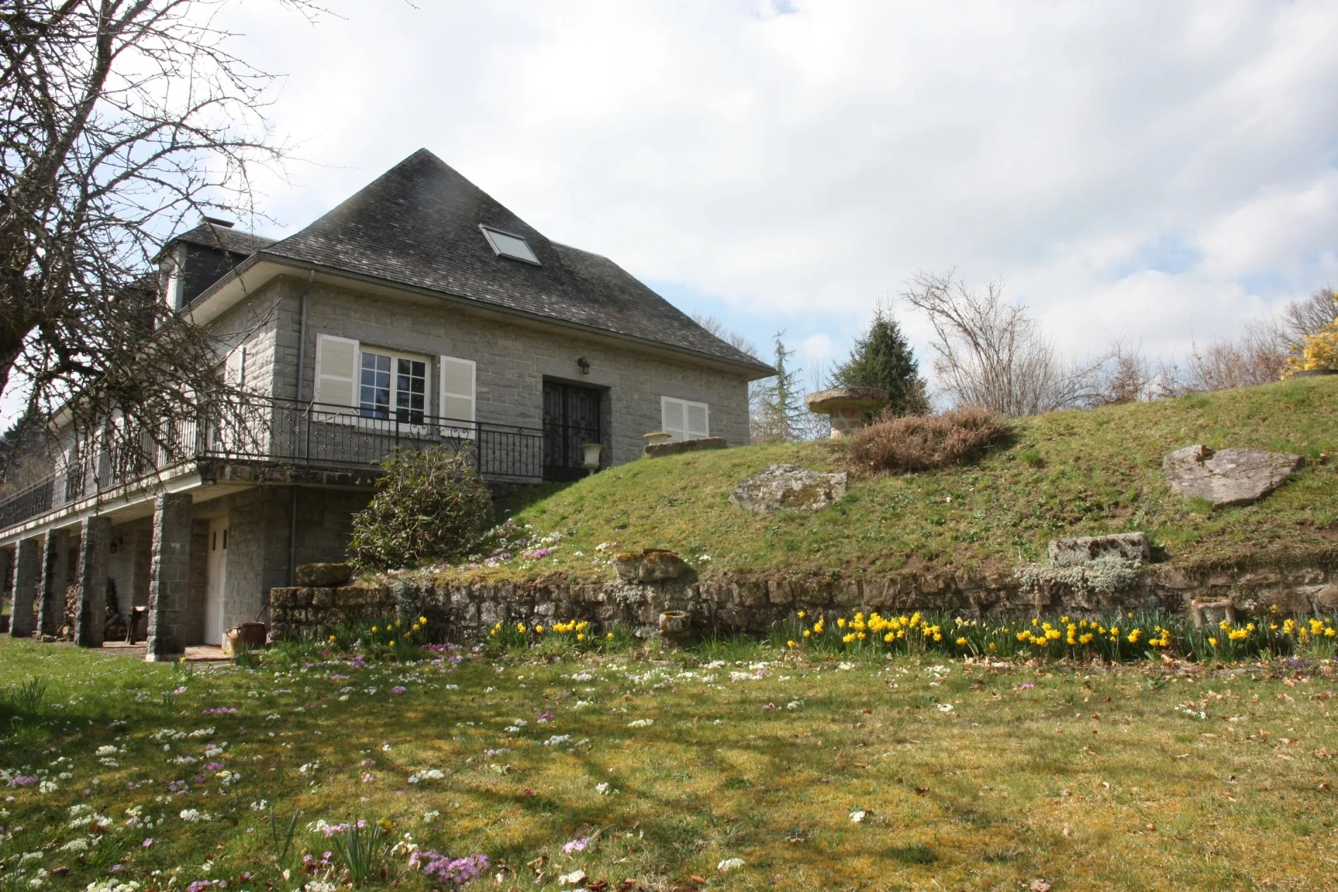 Vue sur la maison depuis le jardin fleuri, avec balcon à l’étage et façade en pierre.