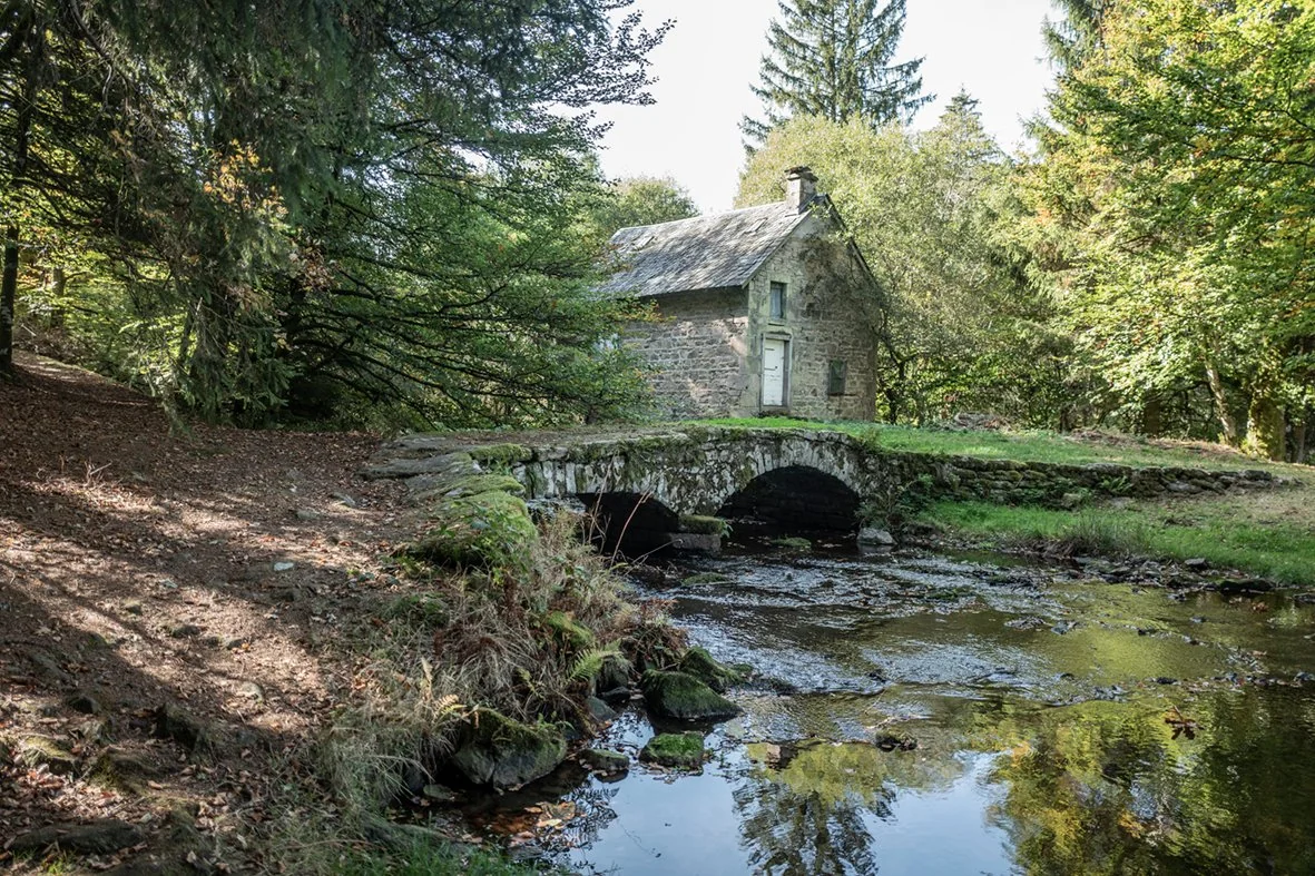 Ancienne maison en pierre au bord d’un ruisseau, traversé par un pont en pierre entouré de forêt.