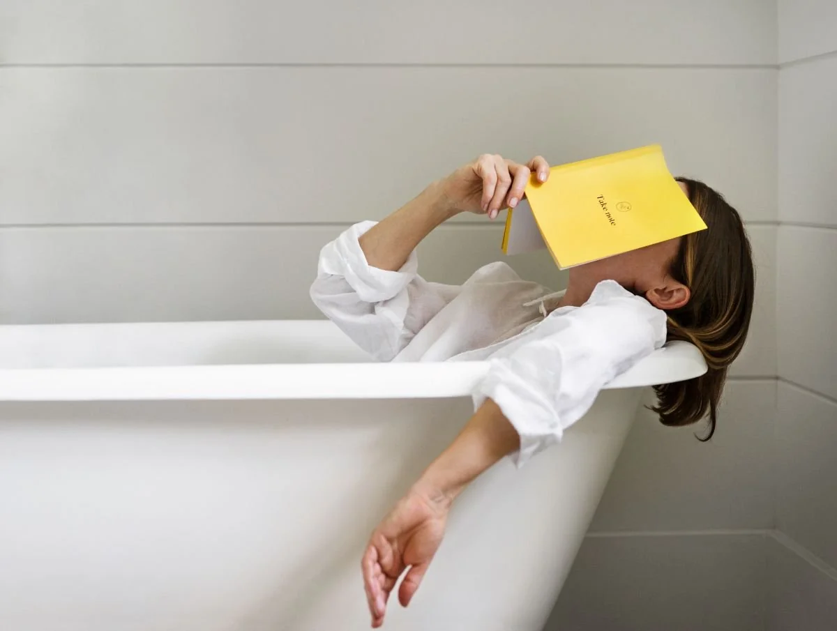 Woman reading a yellow book sitting in a white bathtub