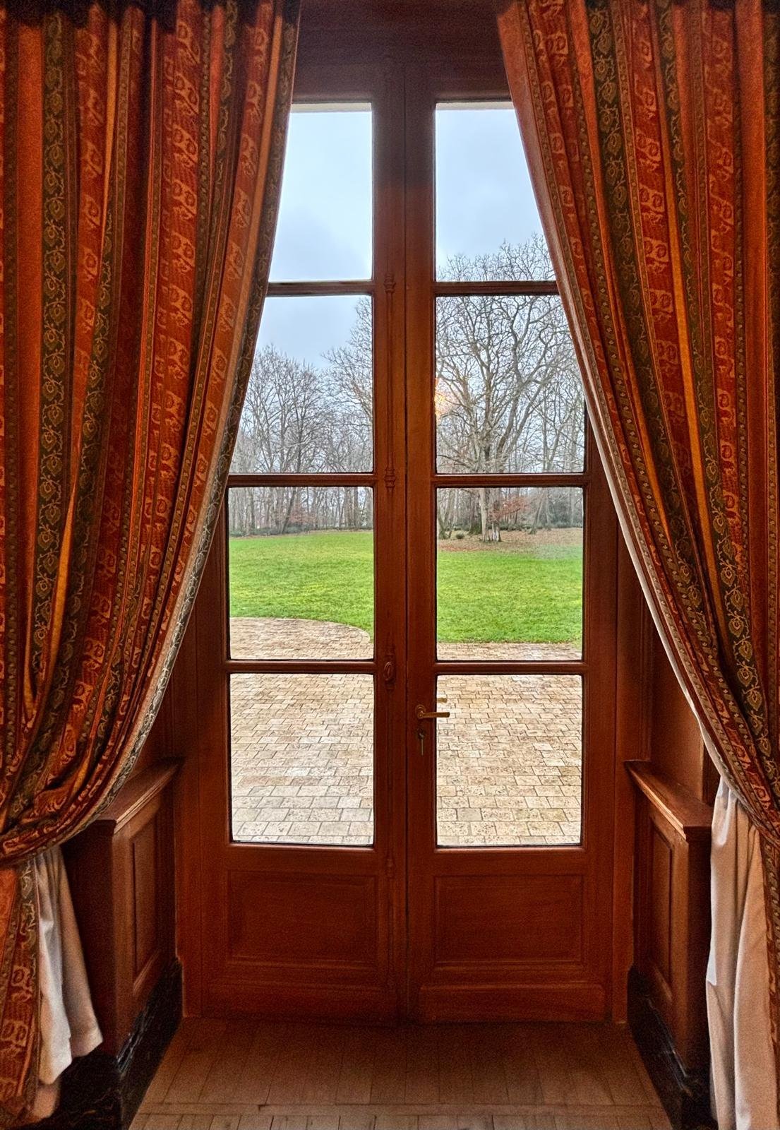 Château de la Rozelle, Vue depuis l'intérieur d'une maison à travers une porte-fenêtre avec des rideaux, donnant sur un jardin.