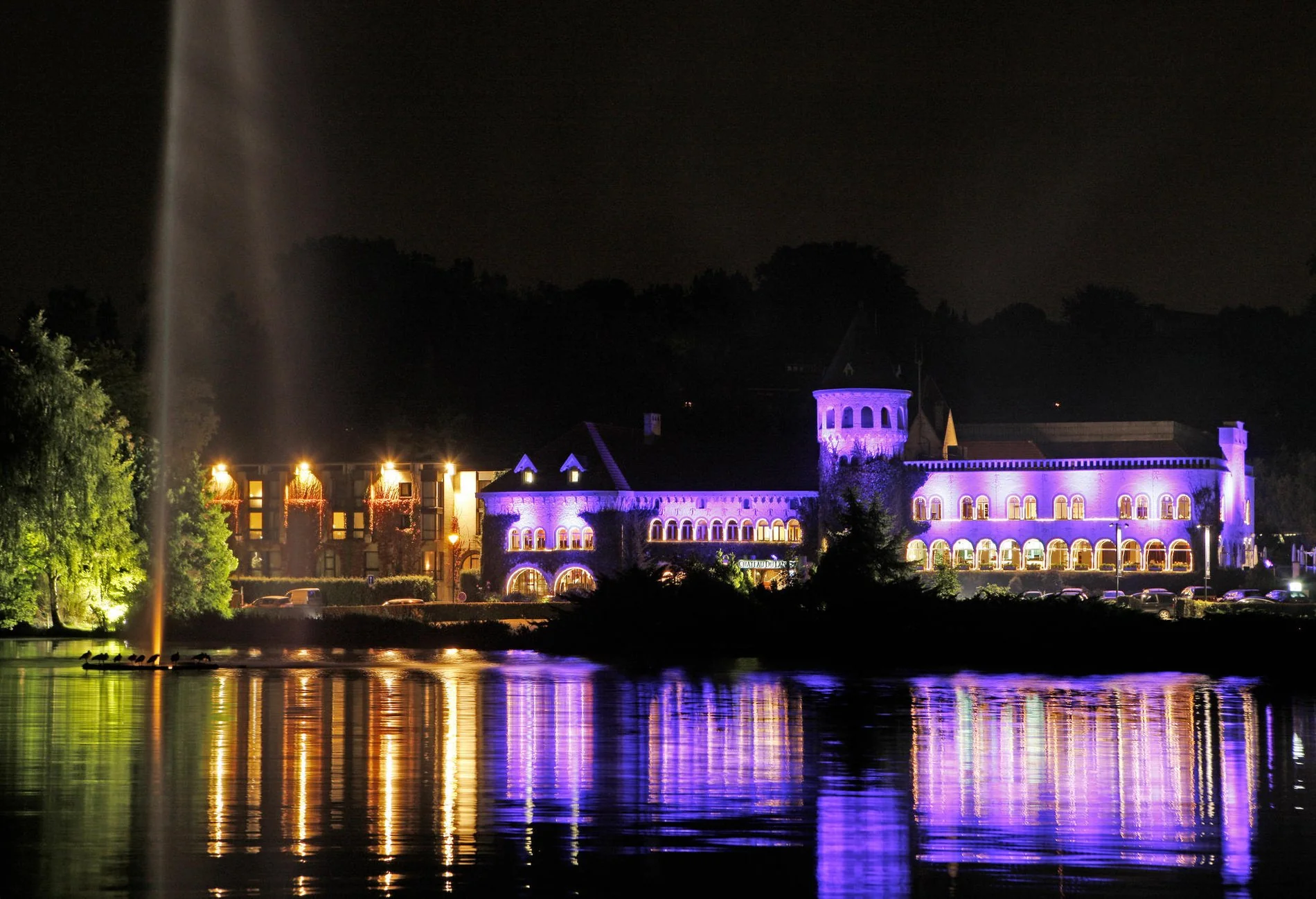 Vue nocturne du Martin's Château du Lac illuminé avec reflets colorés sur le lac.
