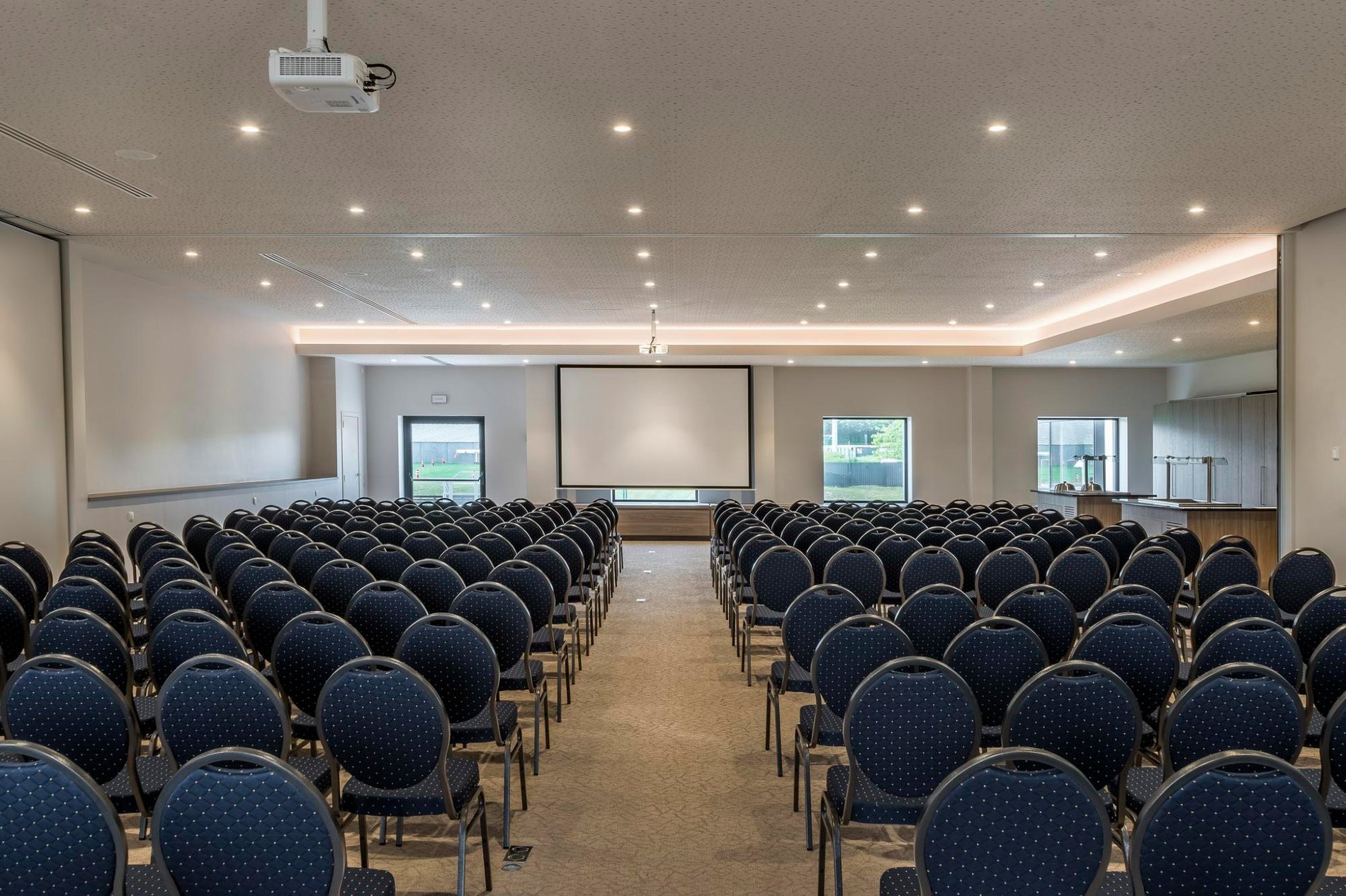 Ruime vergaderzaal met blauwe stoelen op een rij en een projector aan het plafond.